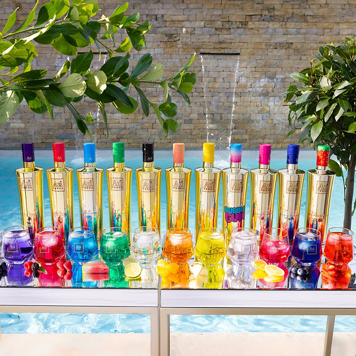 Colorful bottles and glasses on a table by a poolside with plants in the background