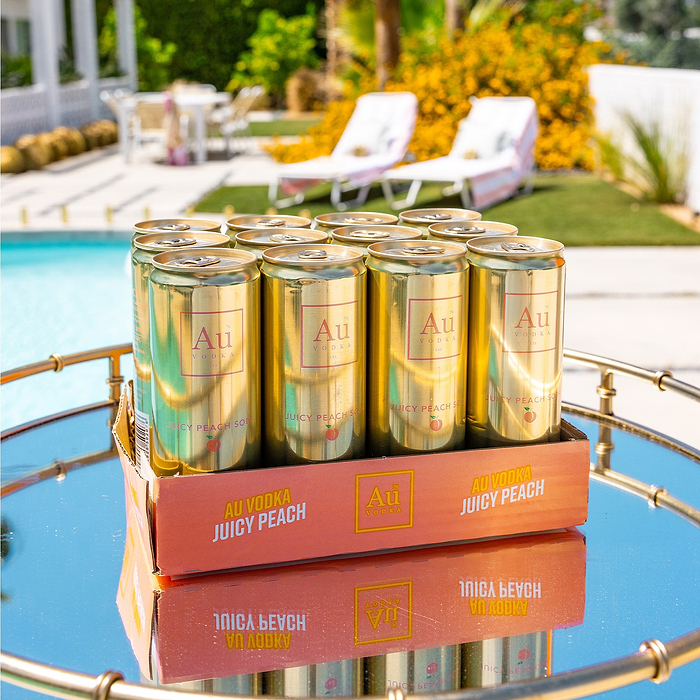 Case of Au Vodka Juicy Peach drinks on a table by a poolside.