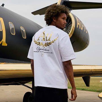 Person wearing a white t-shirt with gold and black design in front of a black and gold airplane.