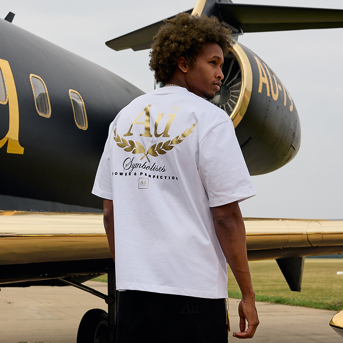 Person wearing a white t-shirt with gold and black design in front of a black and gold airplane.