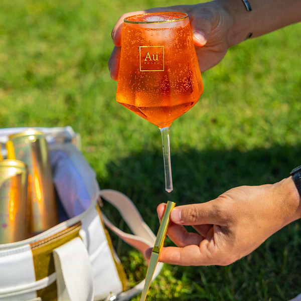 Hand holding an Au Vodka branded glass filled with orange cocktail over a cooler bag