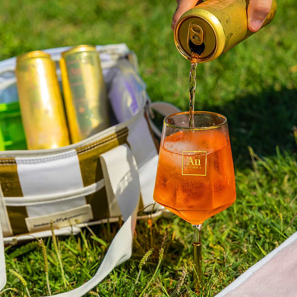 Au Vodka USA canned drink being poured into a glass with ice next to a striped cooler bag on grass