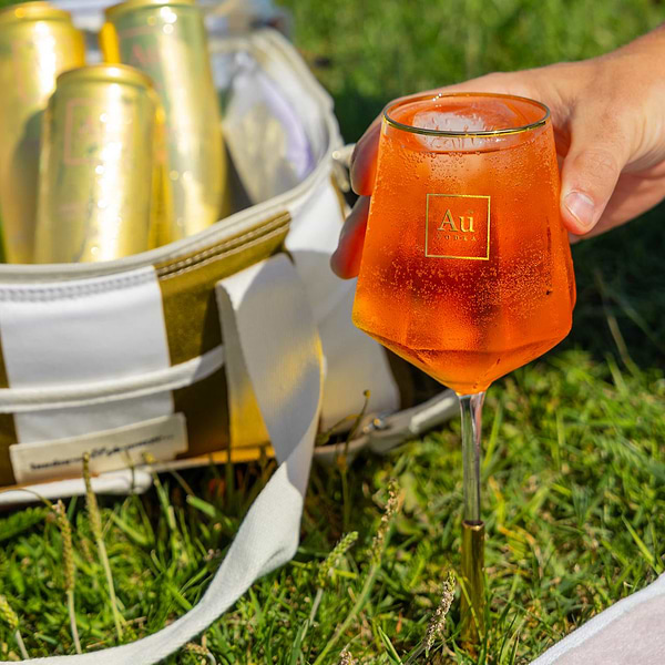 Hand holding an Au Vodka branded glass filled with orange cocktail next to a cooler bag with gold cans on grass