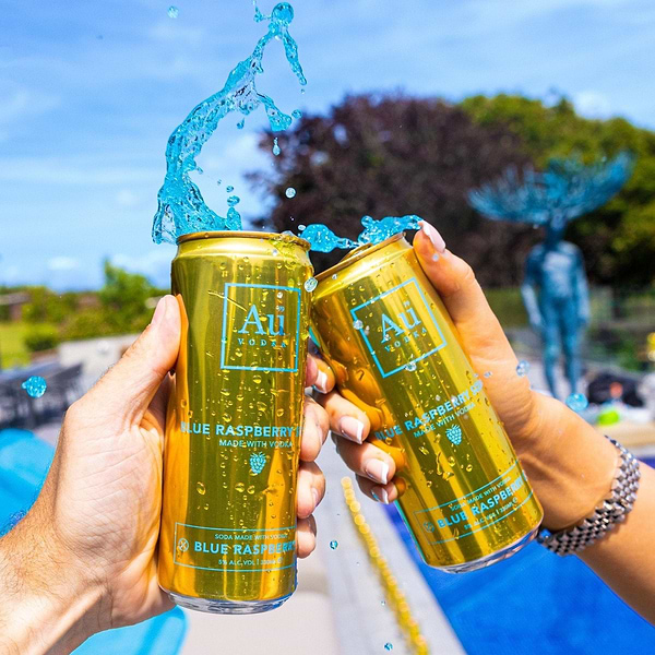 Two hands holding gold Au Vodka Blue Raspberry cocktail cans with blue liquid splashing out
