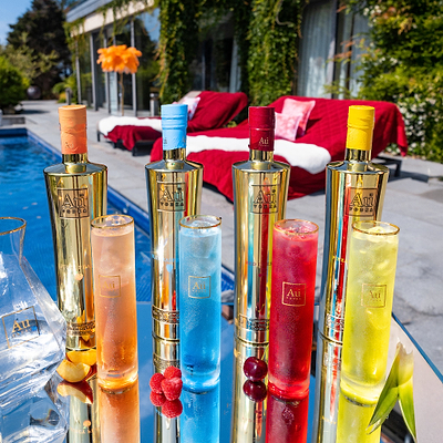 Four Au Vodka bottles in gold with different colored caps, and four matching cocktails on a reflective surface