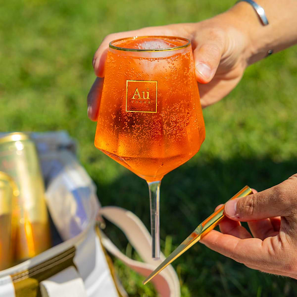 Hand holding an Au Vodka branded glass filled with an orange cocktail and ice outdoors