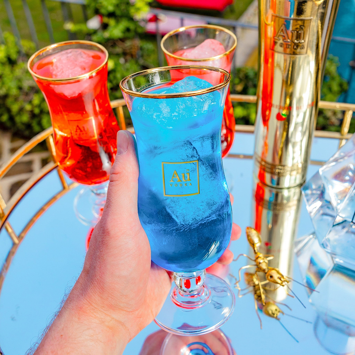 Hand holding an Au Vodka branded glass filled with blue cocktail and ice on a mirrored tray