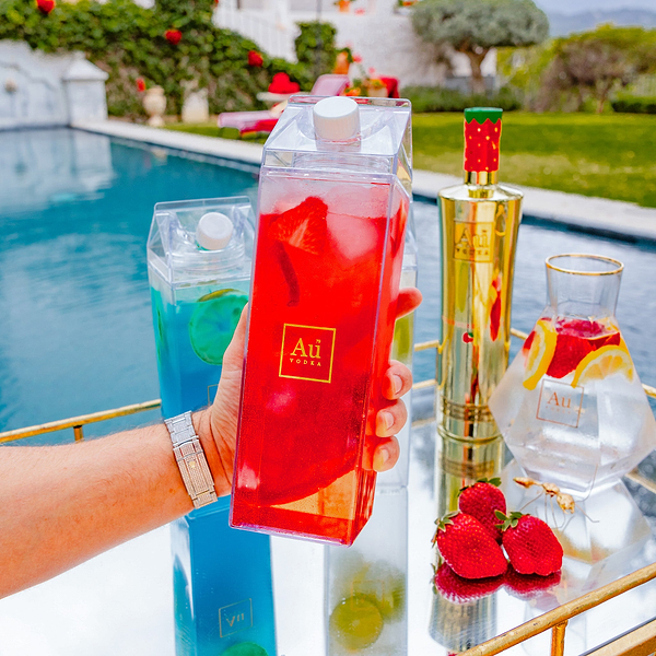 Hand holding a clear carton of red cocktail with ice next to Au Vodka bottle and glass
