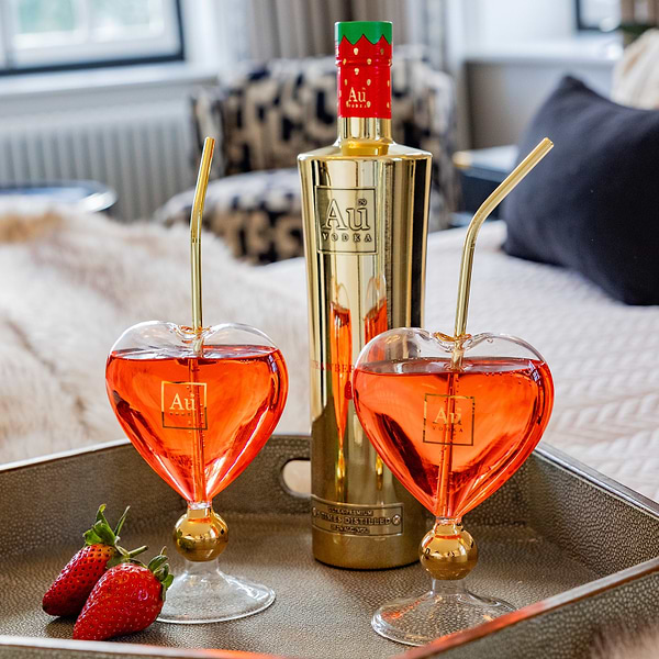 Au Vodka bottle and two heart shaped glasses with red cocktail and gold straws on a tray with fresh strawberries