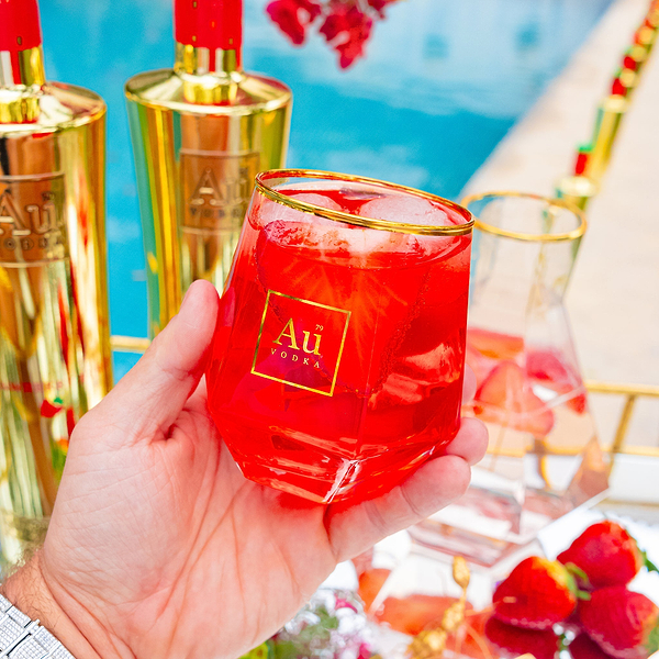 Hand holding a glass of red Au Vodka cocktail with ice next to gold Au Vodka bottles by a pool