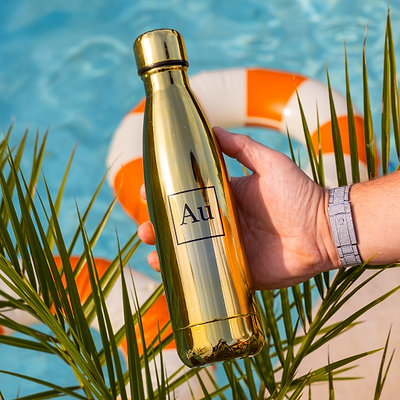 Hand holding a gold Au Vodka USA water bottle with a pool and palm leaves in the background