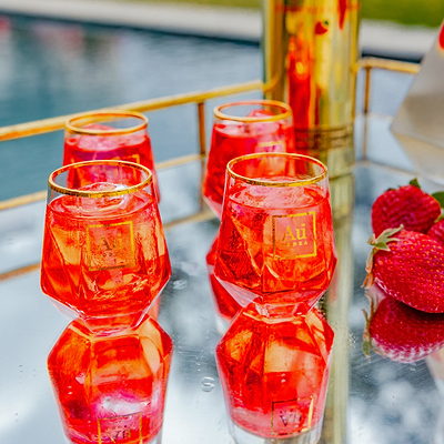Four Au Vodka branded glassware tumblers filled with red liquid and ice on a mirrored tray with fresh strawberries