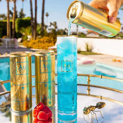 A hand pouring Au Vodka Blue Raspberry Soda into a glass next to two cans and raspberries