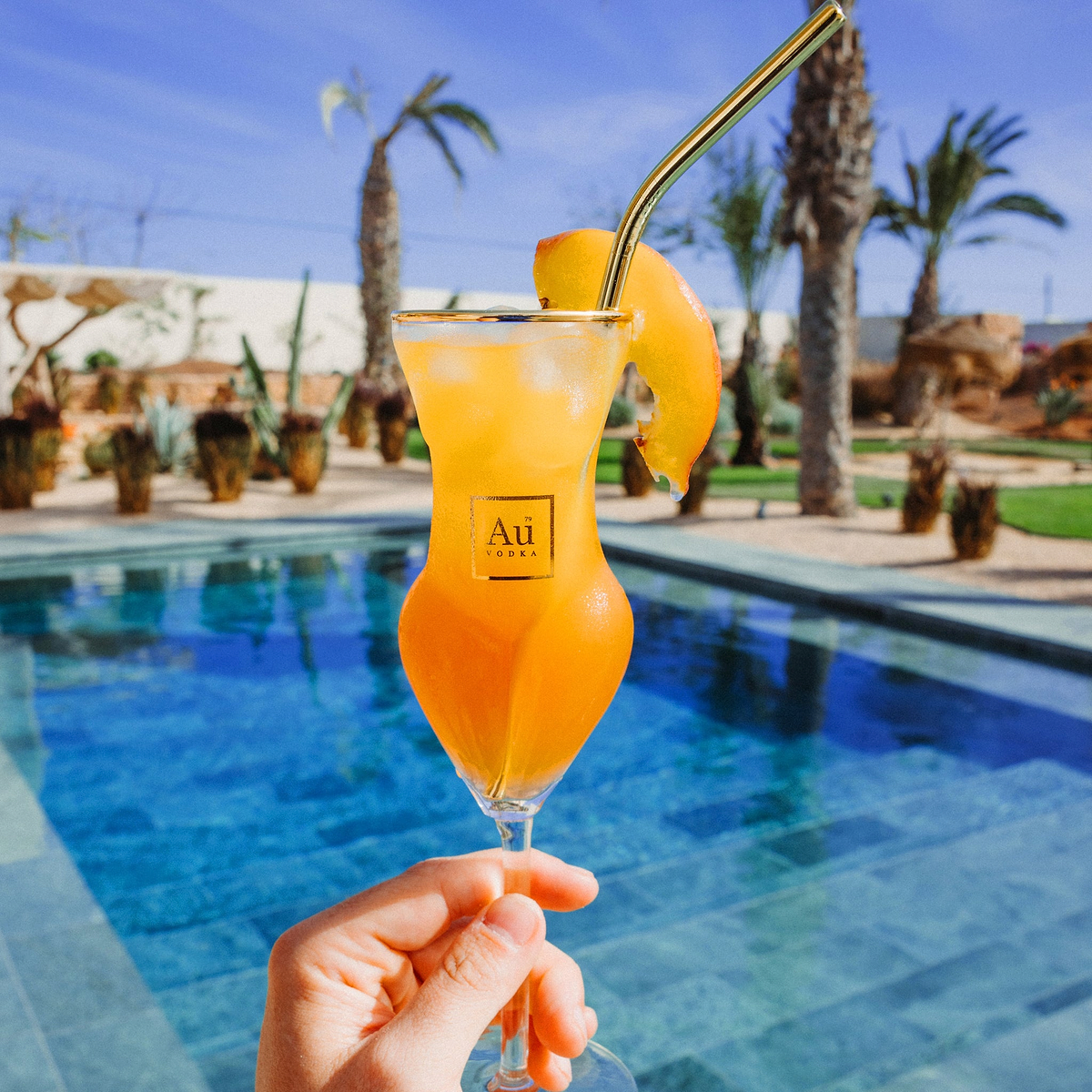 Au Vodka branded glassware filled with orange cocktail and peach slice by a swimming pool