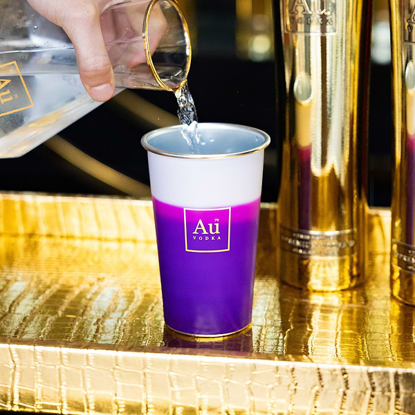 Hand pouring water into a purple and white Au Vodka USA merchandise cup on a gold textured surface
