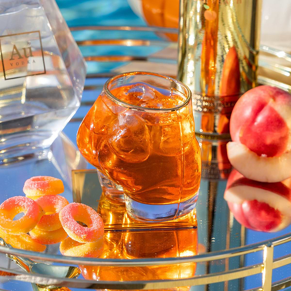 Au Vodka glassware filled with orange cocktail and ice on a mirrored tray with peach slices and candy rings