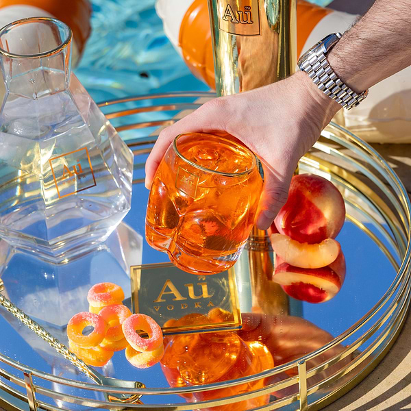 Hand holding an Au Vodka glassware tumbler filled with an orange cocktail and ice on a mirrored tray