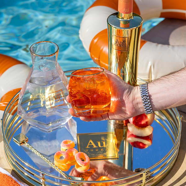 Au Vodka glassware set with a gold bottle and orange cocktail on a mirrored tray by a pool