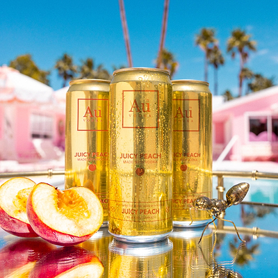 Three Au Vodka USA Juicy Peach cans with peach slices and an ant on a mirrored tray