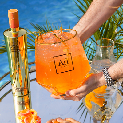 Au Vodka bottle and branded glass filled with orange cocktail on a mirrored tray by a pool