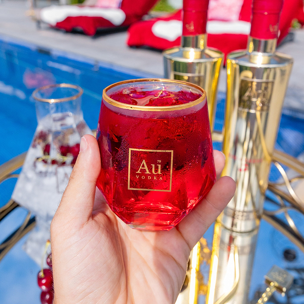Hand holding a glass of red Au Vodka cocktail with gold Au Vodka bottles by a pool