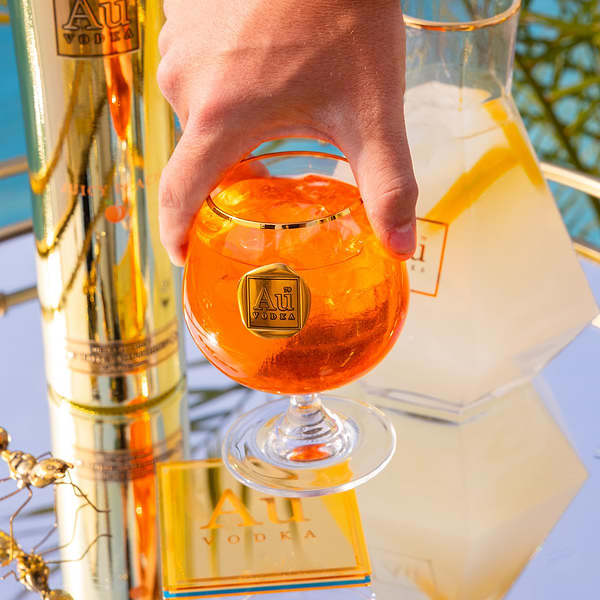 Hand holding an Au Vodka branded glass filled with orange cocktail and ice next to a gold bottle and carafe