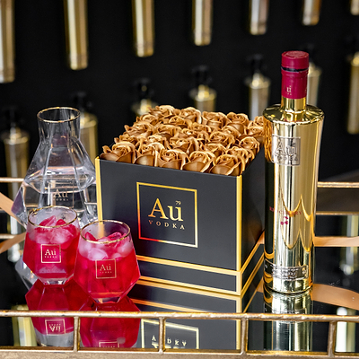 Au Vodka bottle, glasses with red liquid, and a box of gold roses, on a glass table.