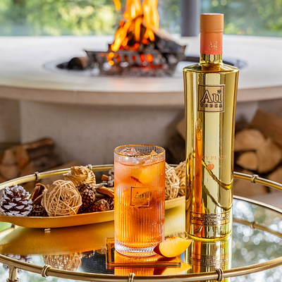 Au Vodka Icy Peach bottle next to a glass of peach cocktail on a gold tray with a fireplace in the background