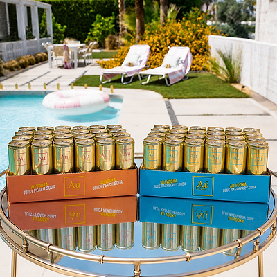 Two 24 packs of Au vodka cocktail cans displayed on a round glass table in front of a poolside patio. The left tray holds juicy peach cans, and the right tray holds blue raspberry cans.