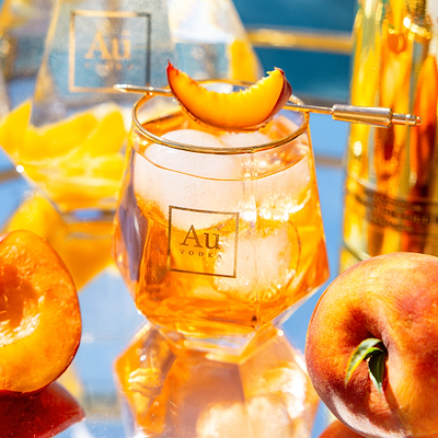 A close up shot of Au Vodka Diamond Cut Glass, filled with Au Vodka Juicy Peach & Ice Cubes, with a peach slice garnish resting on top of the glass. 