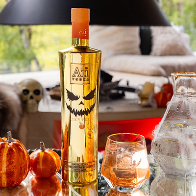 Gold bottle of Au Vodka with a jack-o'-lantern design on a table with pumpkins and a glass.