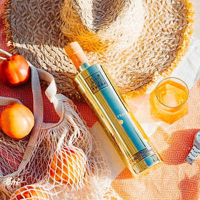 A Bottle of Au Vodka Juicy Peach, sitting on a beach towel, in a summery setting, next to a Au Diamond Cut Glass and other beach items like a Hat, A knitted bag. 