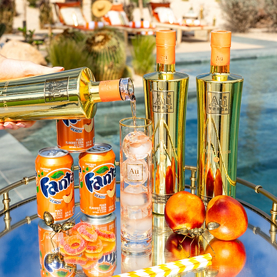 Three Bottles of Au Vodka Juicy Peach, one being poured into an Au Highrise Glass filled with Ice. Placed on a reflective surface, with 3 Cans of Peach Flavored Fanta.