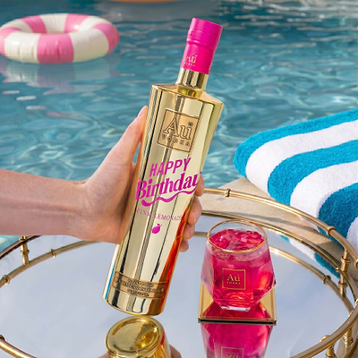 Hand holding a 750ml Au Vodka Happy Birthday Pink Lemonade bottle by a pool with a drink