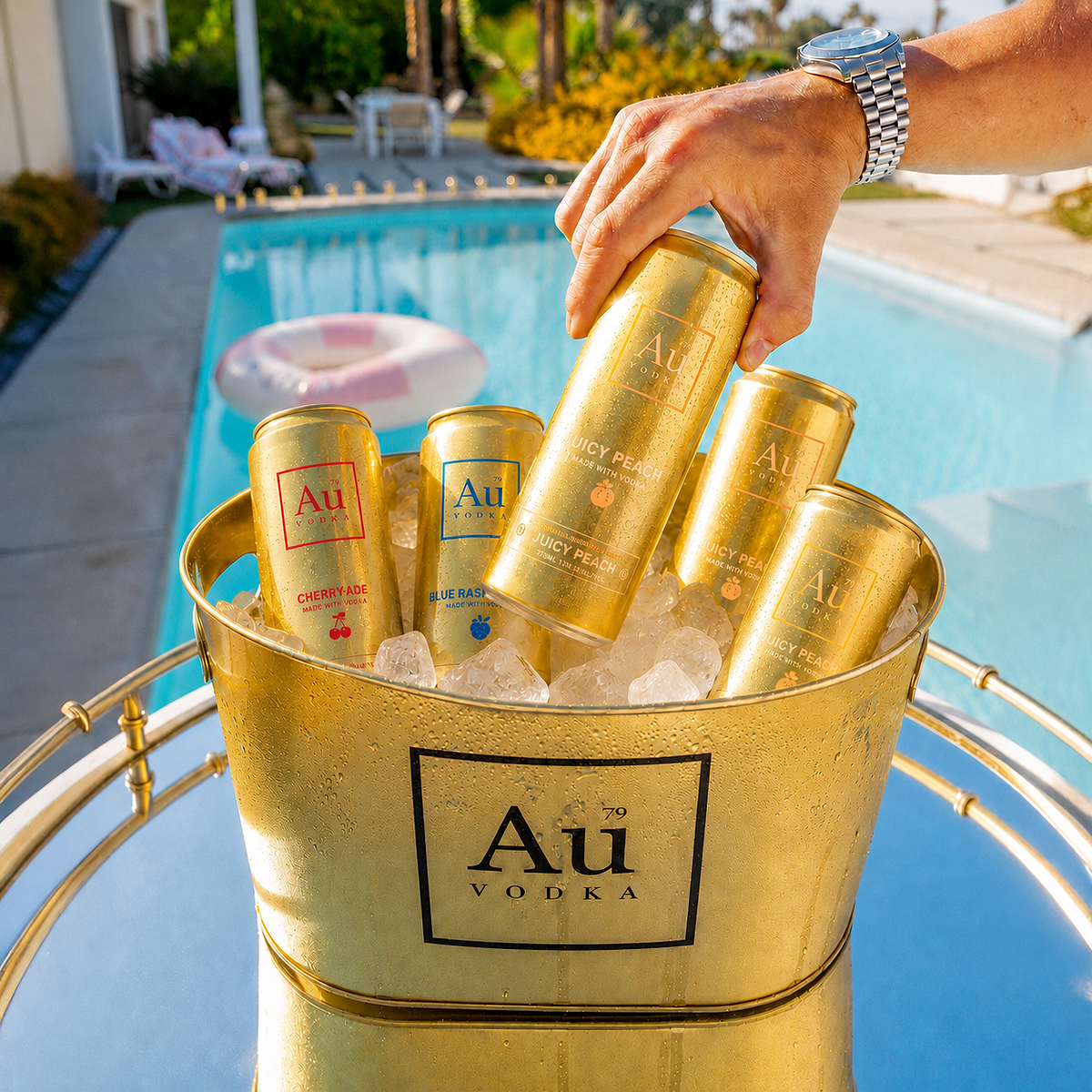 Hand holding an Au Vodka Juicy Peach can over a gold ice bucket filled with assorted Au Vodka cans and ice by a pool