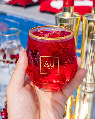 Hand holding a glass of red Au Vodka cocktail with ice near a pool