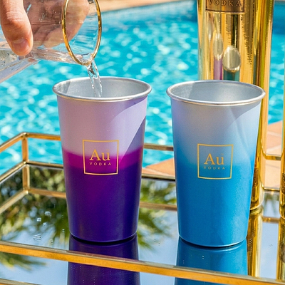 Hand pouring liquid into an Au Vodka USA purple and white ombre cup next to a blue and white cup