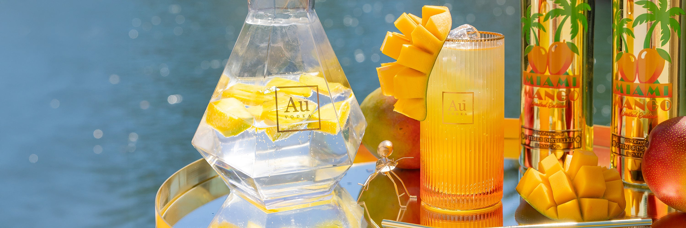 Au Vodka bottle with mango slices, a glass of mango drink, and two Miami Mango vodka bottles
