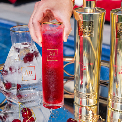 Hand holding an Au Vodka branded highball glass filled with a red cherry cocktail next to Au Vodka bottles