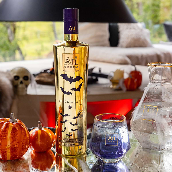 Au Vodka Black Grape bottle with bat graphics next to a glass of purple liquid and Halloween pumpkins