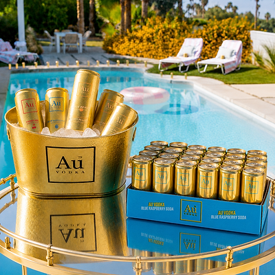 A gold bucket and matching tray display Au Vodka Blue Raspberry Soda cans in an outdoor poolside setting with lounge chairs and palm trees in the background.