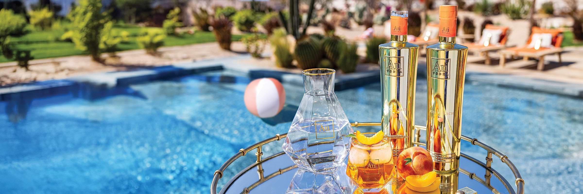 Two gold Au Vodka Juicy Peach bottles and a cocktail on a mirrored tray by a swimming pool