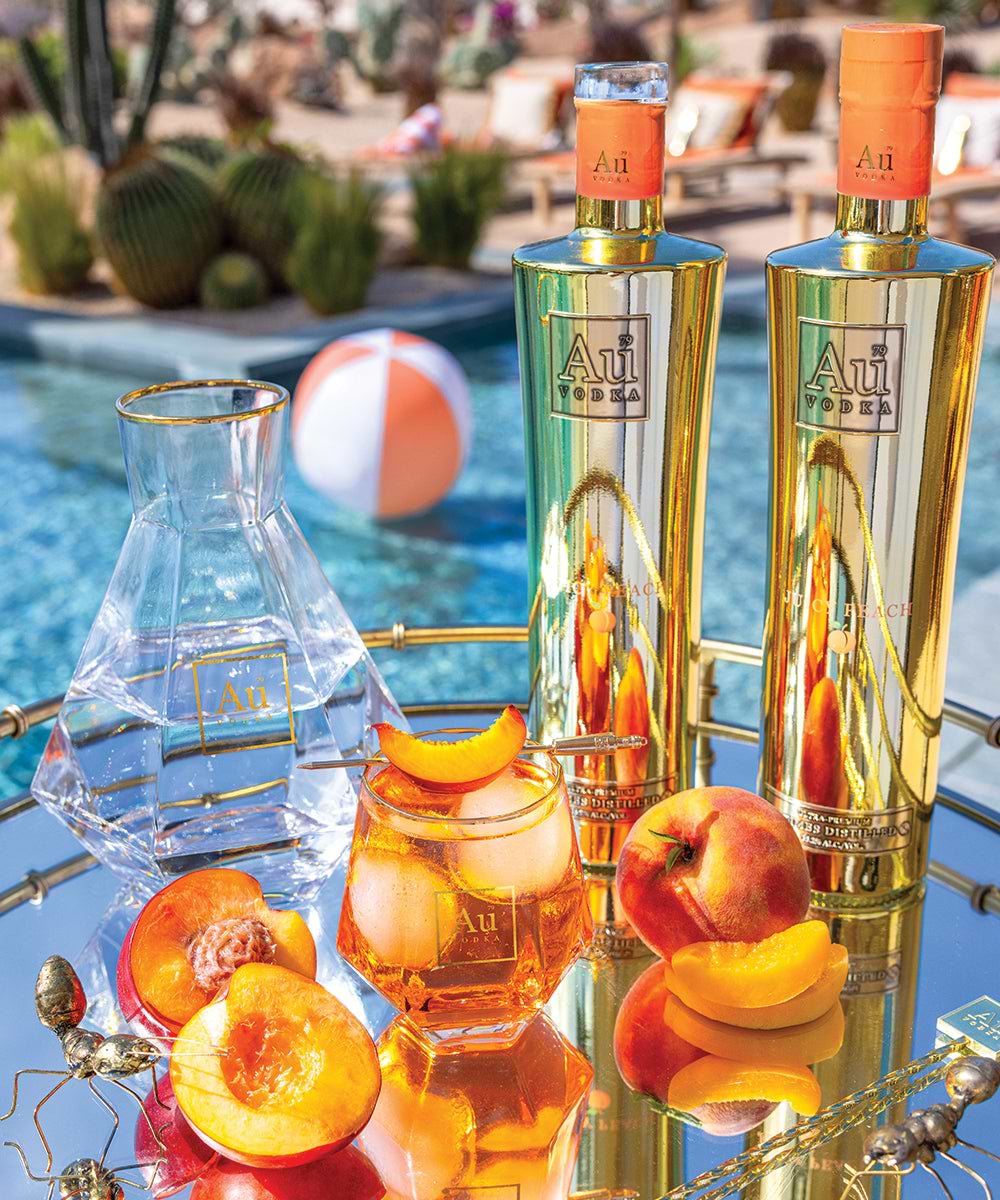 Two gold Au Vodka Juicy Peach bottles with a peach cocktail and fresh peach slices on a mirrored tray by a pool