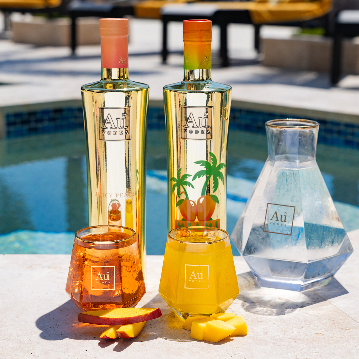 Au Vodka Juicy Peach and Miami bottles, two glasses of cocktails, and a decanter by a pool