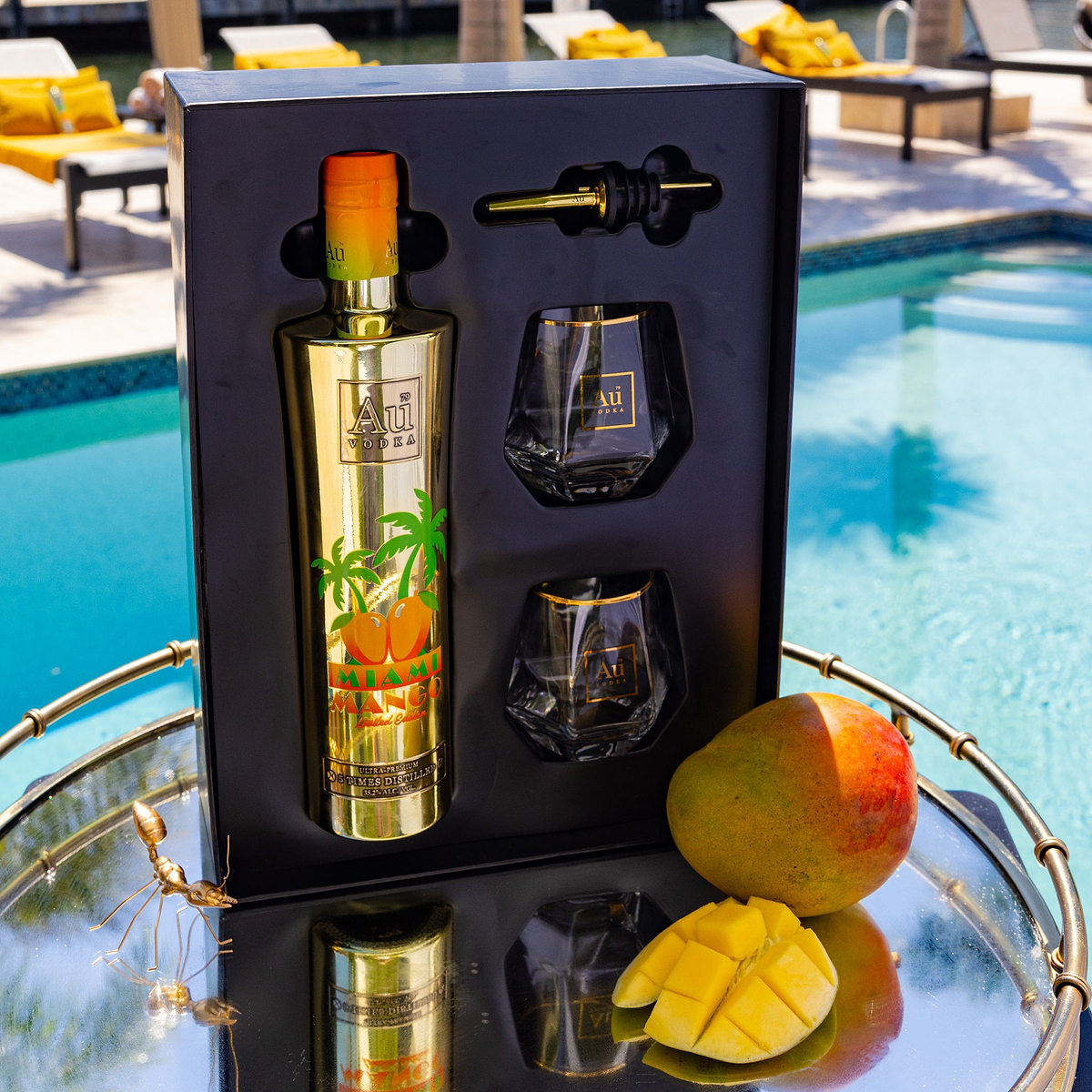 Au Vodka Miami Mango Gift Pack with bottle, two glasses, and pourer, next to whole and sliced mango