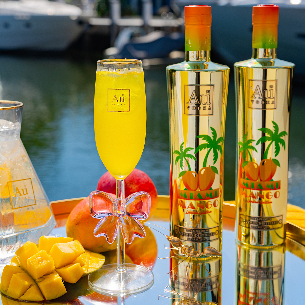 Au Vodka Miami Mango Bottle & Glassware Bundle with mangoes and a drink on a reflective surface