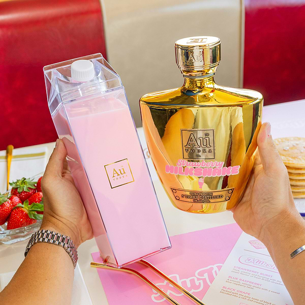 Hands holding a clear carton of pink cocktail and a gold Au Vodka bottle