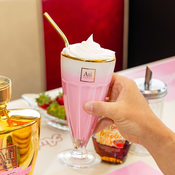 Hand holding an Au Vodka branded pink milkshake with whipped cream and a gold straw