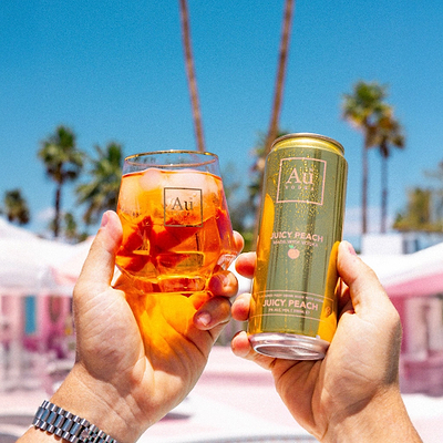 Hands holding an Au Vodka USA Juicy Peach can and a short glass with ice and drink