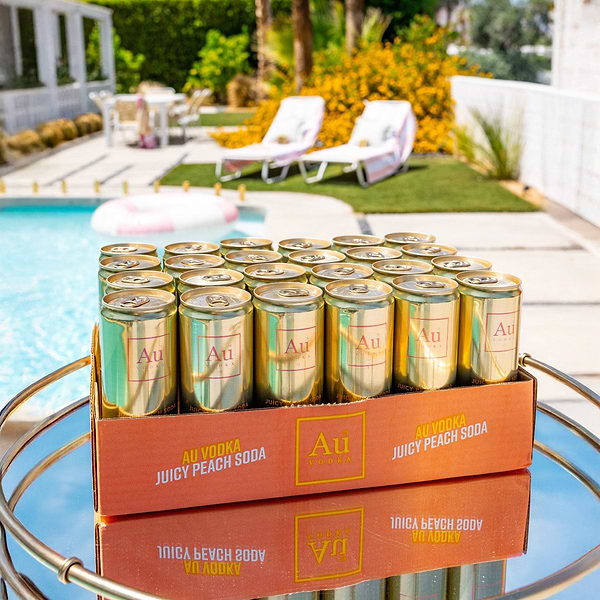 Case of Au Vodka Juicy Peach Soda cans on a mirrored table by a swimming pool